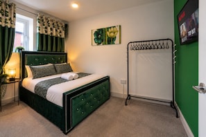 A neatly arranged bedroom with a bed, a bedside table with a lamp, a wall-mounted TV, and a green wall. - Cosy Private House with Hot Tub (Leeds)