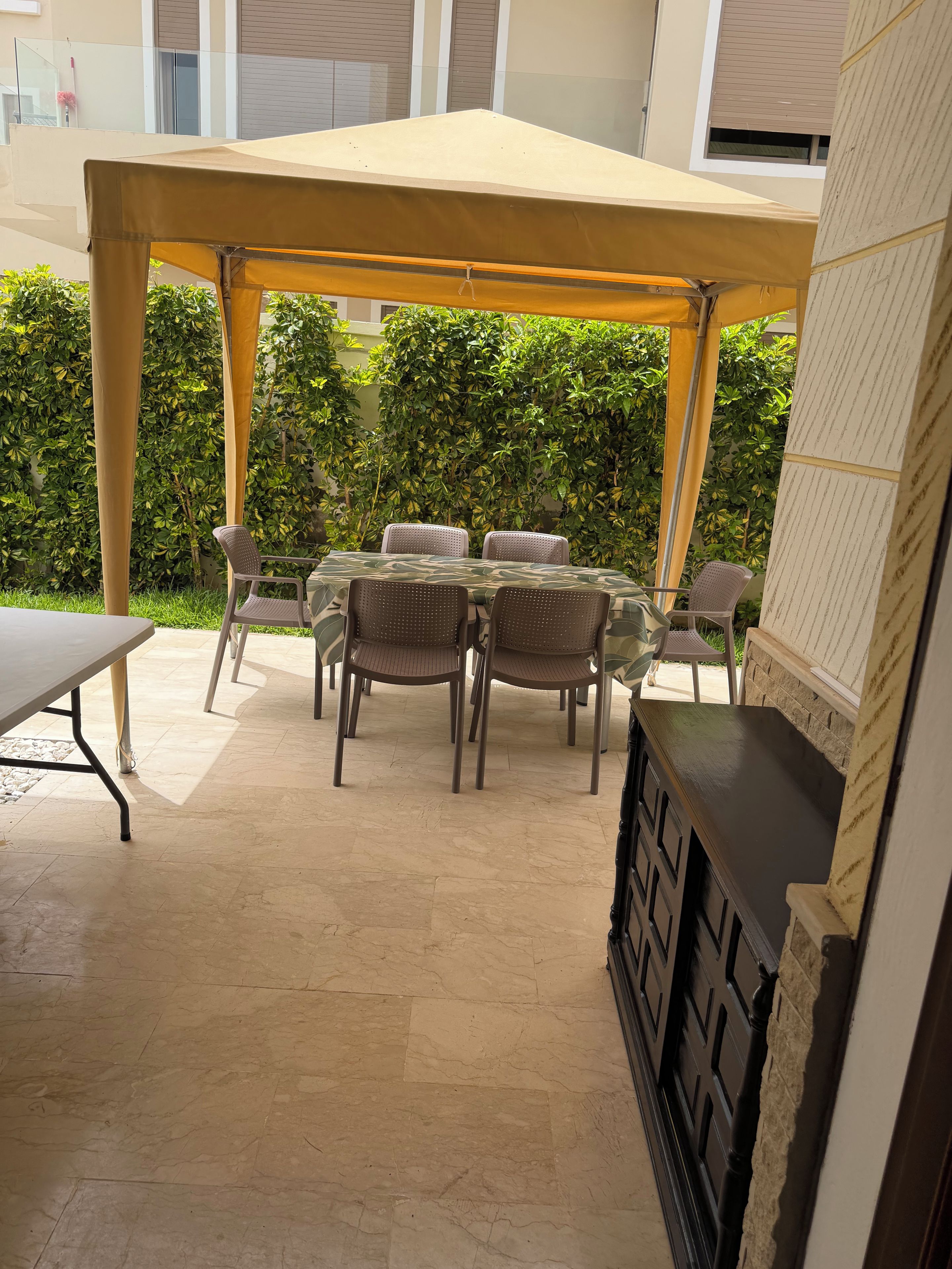An outdoor patio with a covered table and chairs, a fireplace, and a hedge in the background.