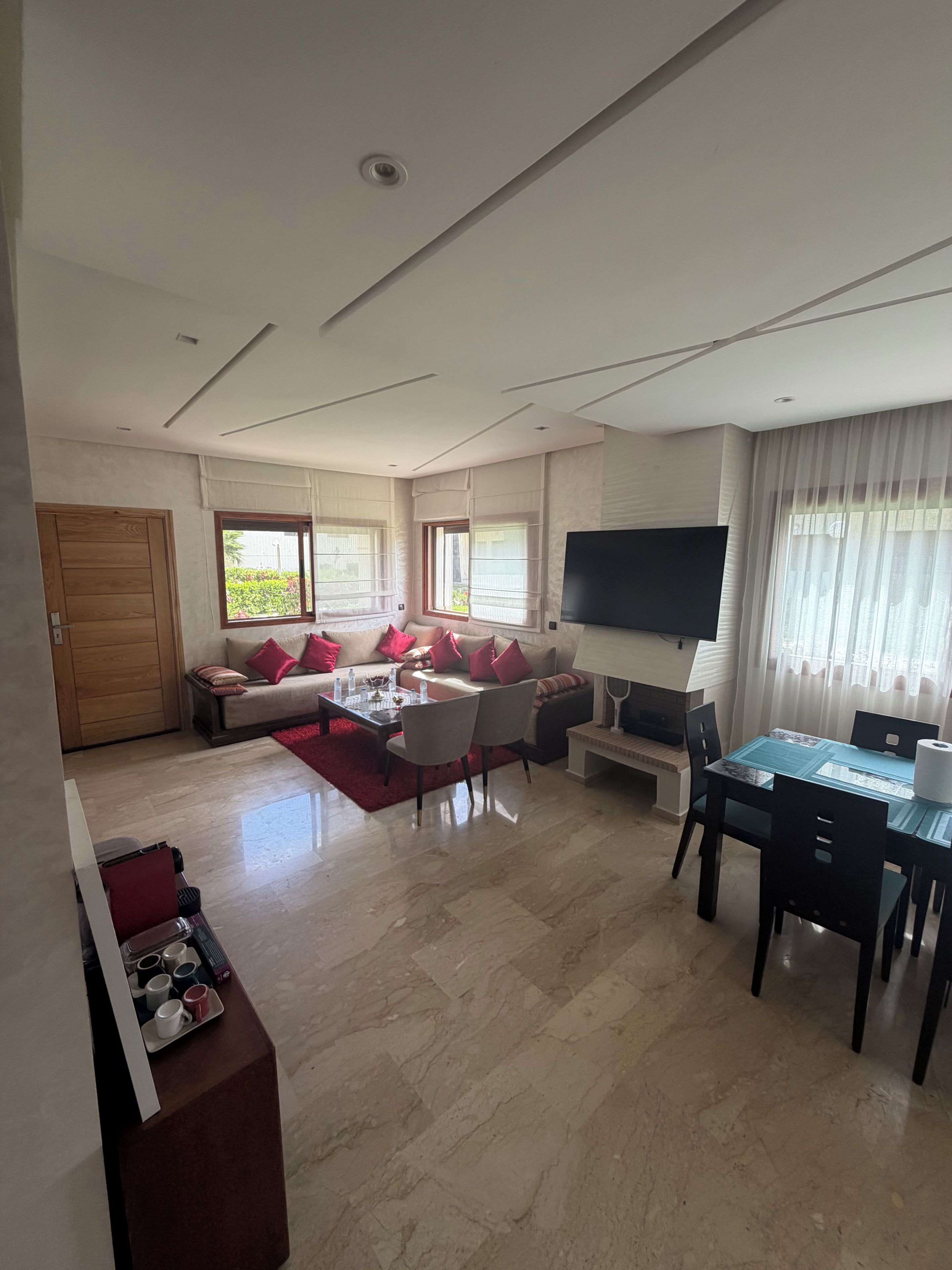 Ein modernes Wohnzimmer mit einer Sofagarnitur, einem Esstisch mit Stühlen, einem an der Wand befestigten Flachbildfernseher und einem großen Fenster mit Vorhängen.