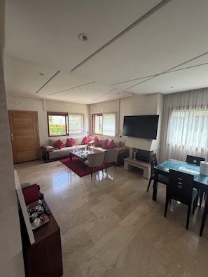 A modern living room with a sofa set, a dining table with chairs, a flat-screen TV mounted on the wall, and a large window with curtains.