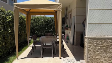 Un espace extérieur abrité, avec une table et des chaises, situé à côté d’un bâtiment dont la façade est en pierre.