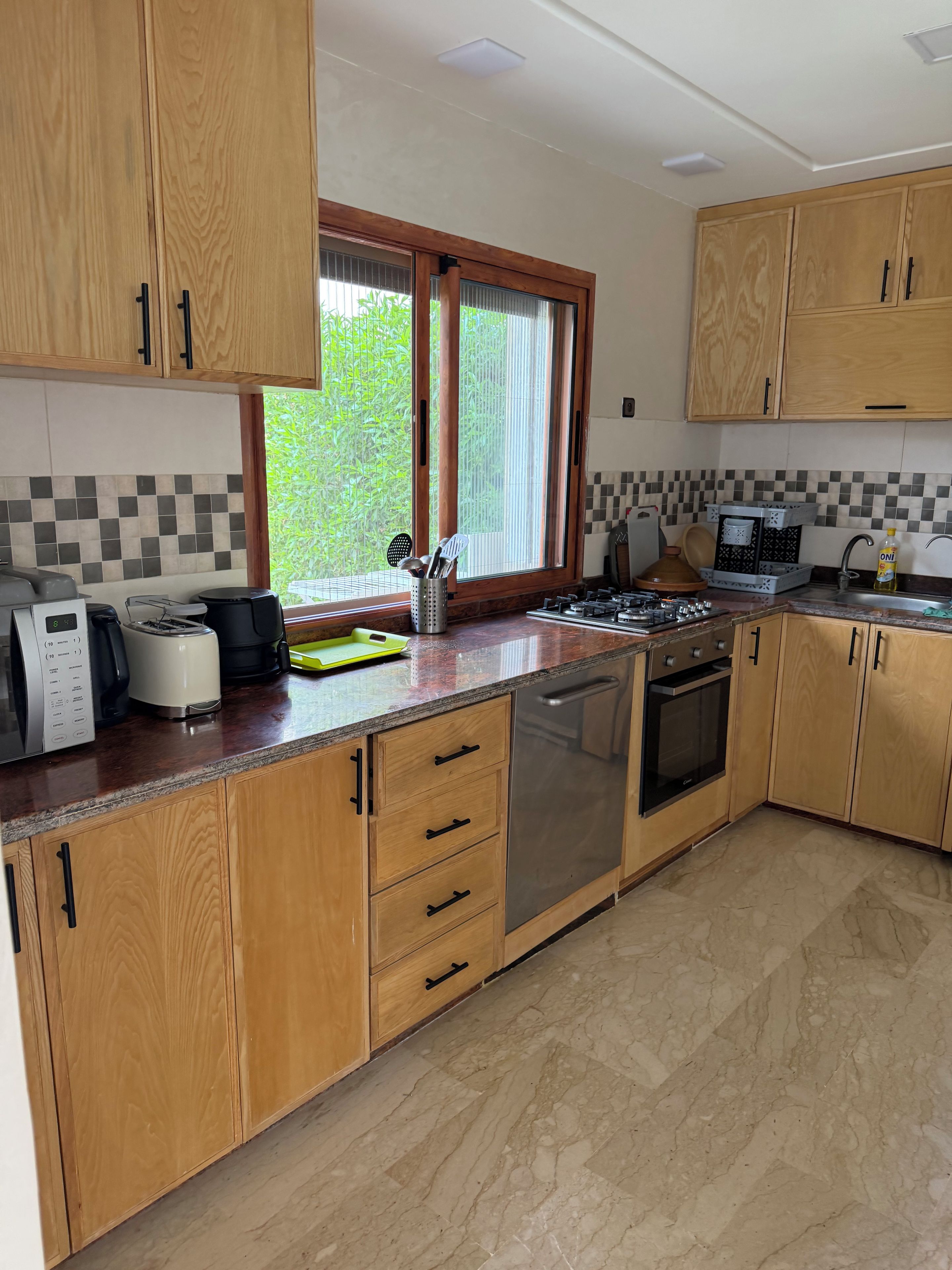Une cuisine avec des meubles en bois, un lave-vaisselle en acier inoxydable, une cuisinière à gaz et une fenêtre donnant sur un paysage verdoyant.