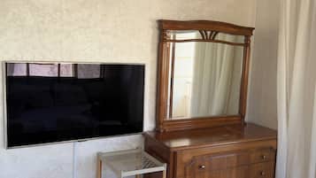 Ein Zimmer mit einem hölzernen Schminktisch, einem an der Wand befestigten Fernseher und einer kleinen Regalwand.