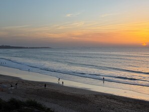 A beach with people walking along the shore and a sunset in the background.