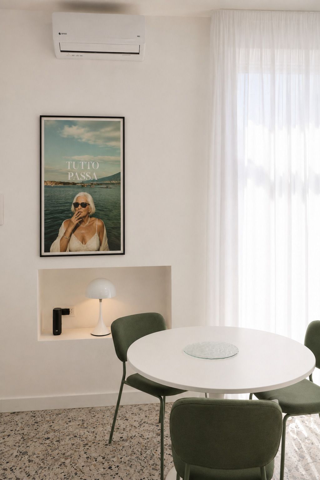 Une chambre d’hôtel avec une table ronde, des chaises vertes, un tableau encadré au mur, une lampe et un appareil de climatisation.