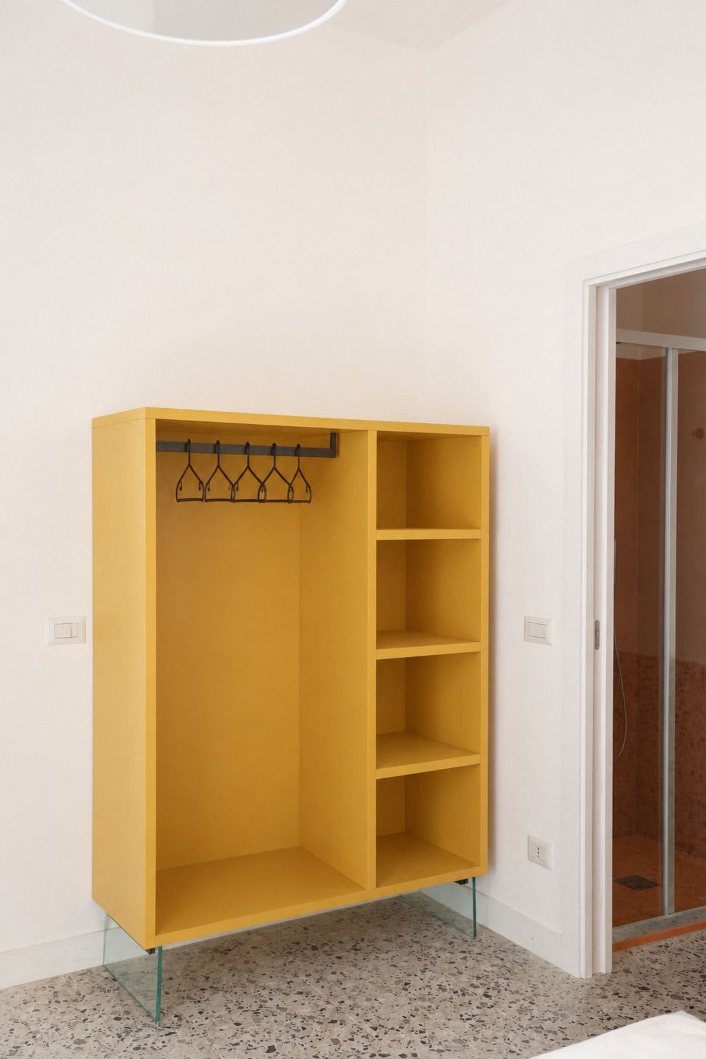 Un armario amarillo con varios estantes y una barra para colgar ropa, situado frente a una pared blanca con una puerta a la derecha.