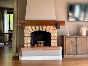Una chimenea con arco de piedra, repisa y base blanca. A la derecha, un mueble de madera y un televisor de pantalla plana en la pared.