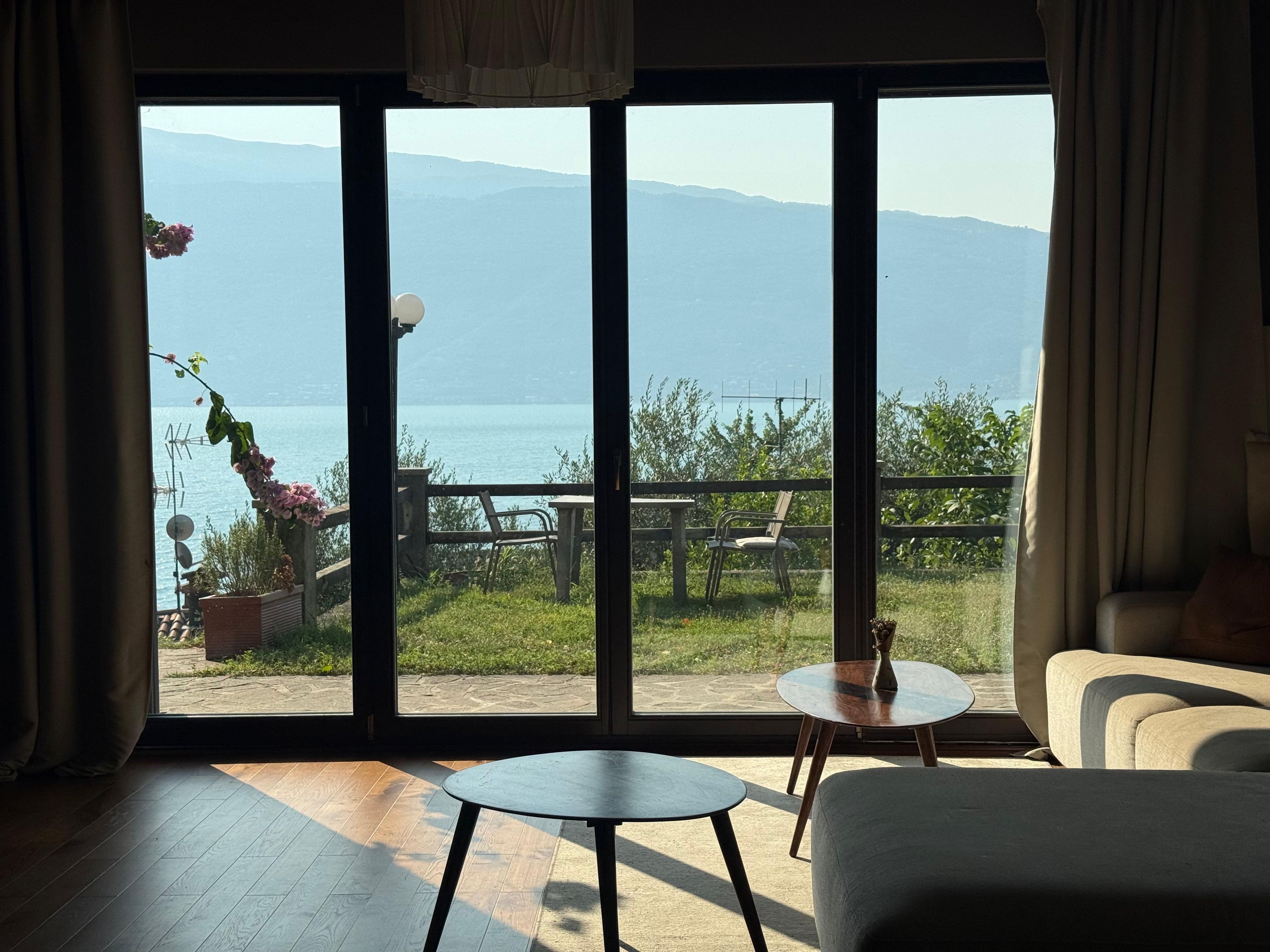 Une pièce dotée d’une grande porte-fenêtre coulissante qui donne sur un espace extérieur offrant une vue sur un plan d’eau et des montagnes. On y trouve deux petites tables rondes et un canapé.