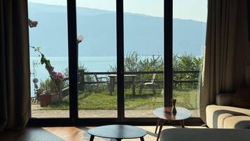 Une pièce dotée d’une grande porte-fenêtre coulissante qui donne sur un espace extérieur offrant une vue sur un plan d’eau et des montagnes. On y trouve deux petites tables rondes et un canapé.