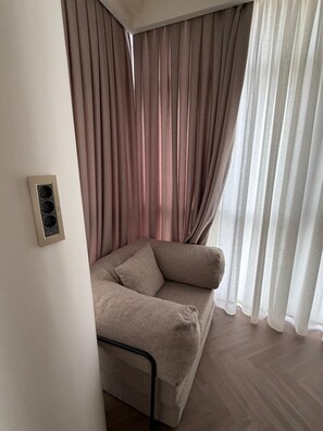 Ein Eckzimmer mit einem beigen Sofa, einer Wandsteckdose und durchsichtigen Vorhängen.