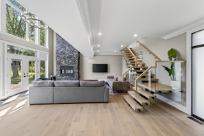 Un salon moderne avec une cheminée en pierre, un canapé d’angle gris et un escalier en bois.