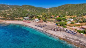 Una zona costera con playa, aguas azules cristalinas y un paisaje montañoso con aerogeneradores al fondo.