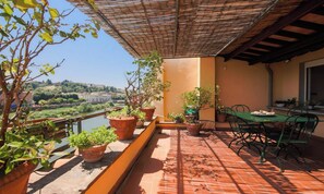 Terraço com mesa, cadeiras, plantas em vasos e vista para um rio e horizonte urbano.