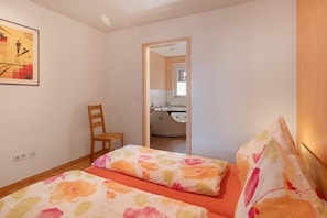 Quarto com duas camas, uma cadeira, um quadro e banheiro visível por uma porta aberta.