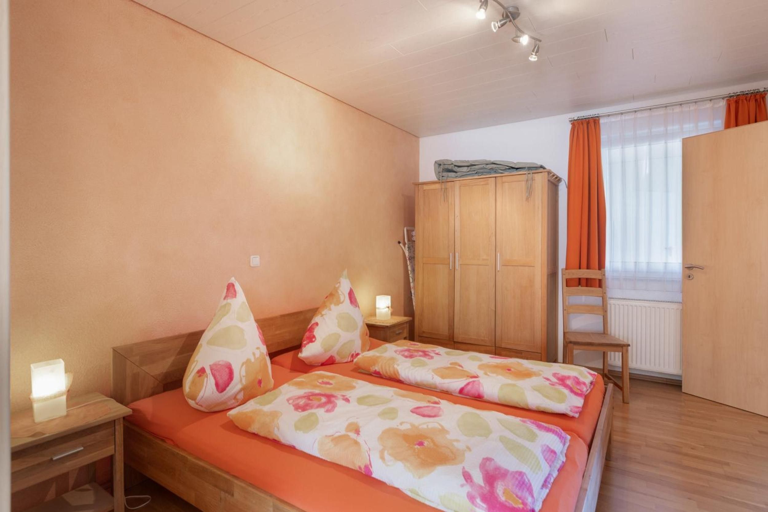A bedroom with a double bed, a wooden wardrobe, a chair, and a window with curtains.