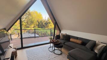 Ein modernes Schlafzimmer im Loft-Stil mit einem großen Dachfenster, einer Sitzecke, einem kleinen runden Tisch und Blick auf eine Terrasse und Bäume.
