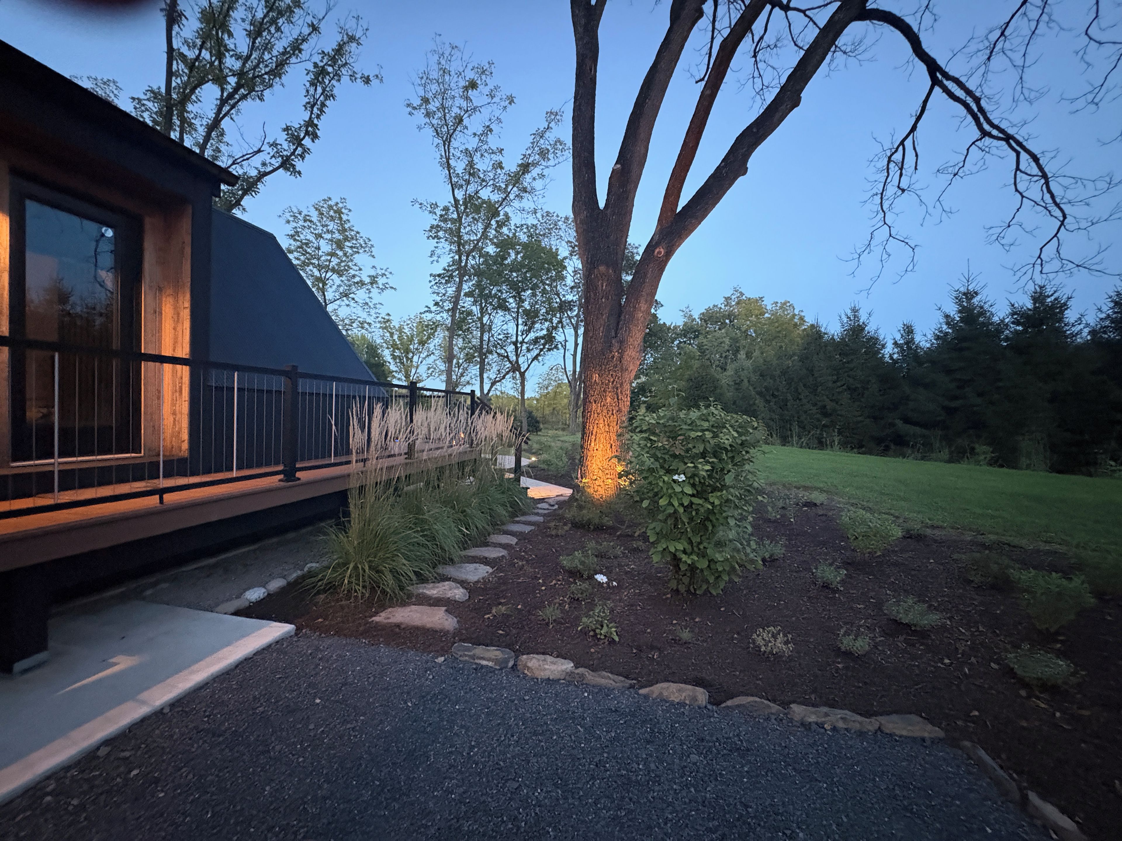 A modern building with a dark roof and large windows, surrounded by a landscaped garden with a stone pathway and a tree.