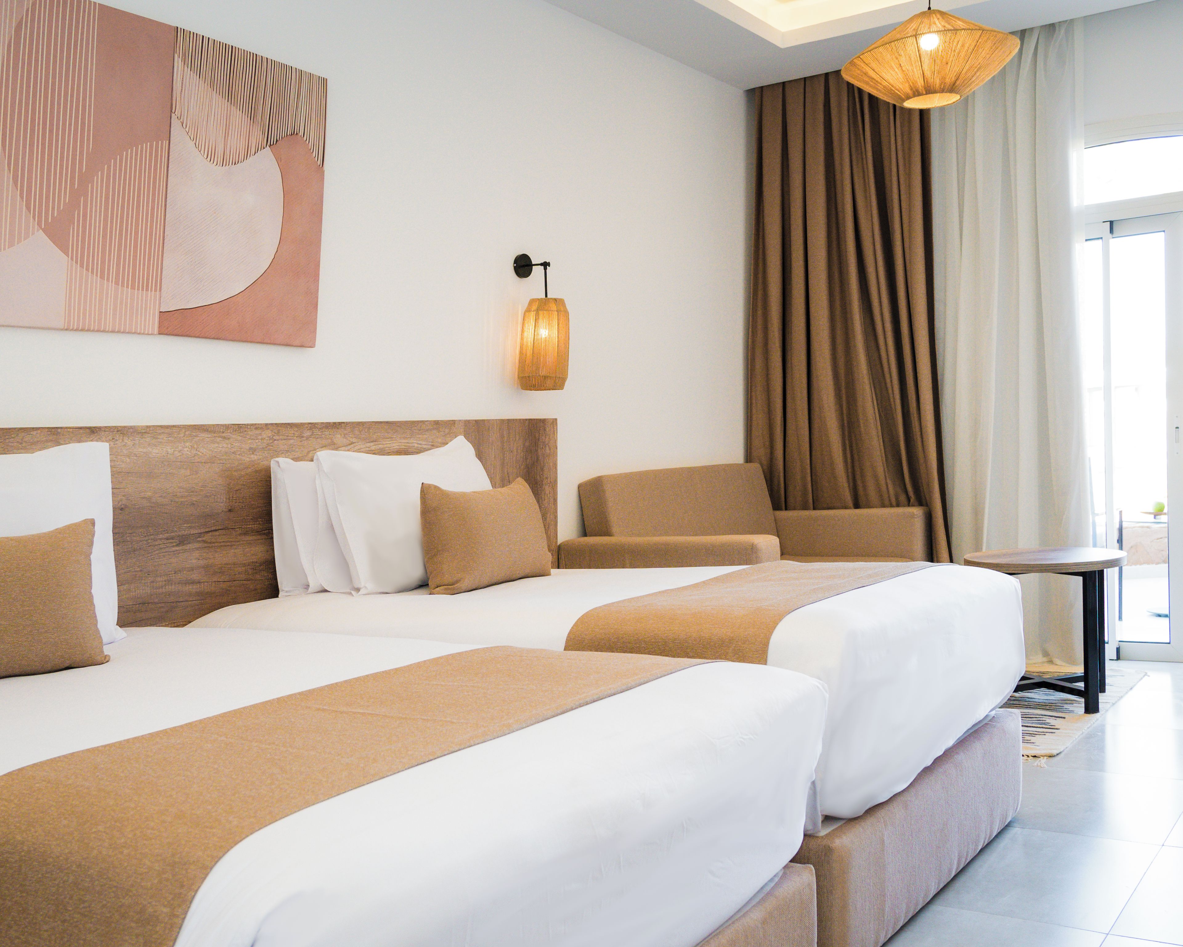 A hotel room with two beds, a wooden headboard, a wall-mounted light fixture, and a view of the outdoors through a window with curtains.