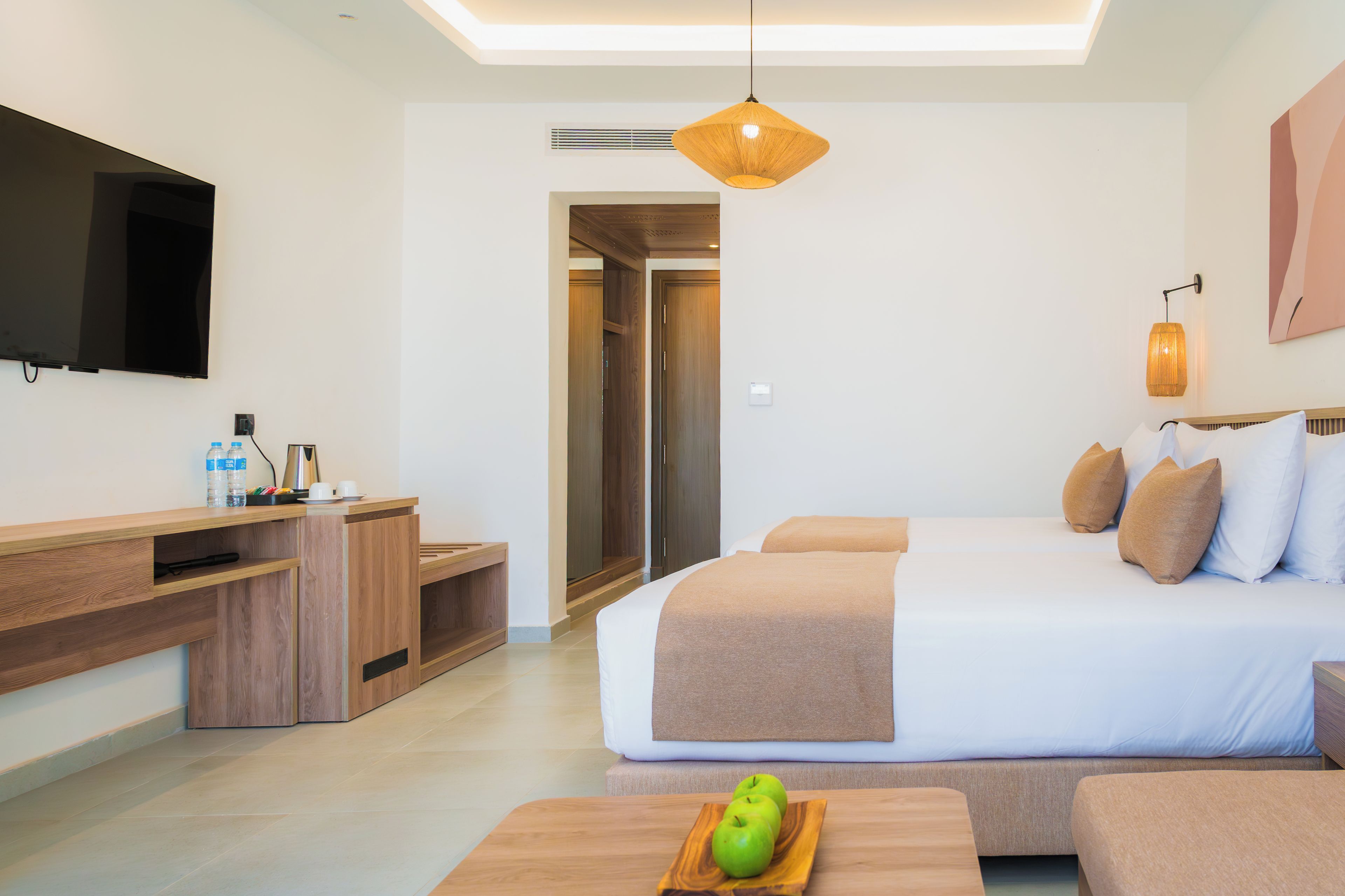 A modern hotel room with a large bed, a desk, a flat-screen TV, and a dining table with apples.