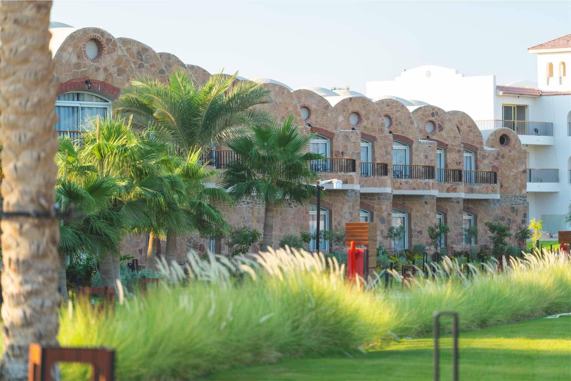 Un hotel de varios pisos con fachada de piedra y balcones, rodeado de palmeras y césped bien cuidado.