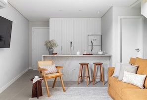 A compact kitchen with a white countertop, a built-in oven, and a refrigerator. There is a wooden chair and two stools in front of the counter. - Duna Loft overlooking the Lagoon | Pool and gym (Florianópolis)