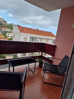 Uma varanda com mesa e cadeiras, um vaso de planta e vista para prédios ao fundo.