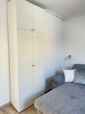 Ein Schlafzimmer mit grauem Sofa, einem weißen Kleiderschrank und einer Wandleuchte.