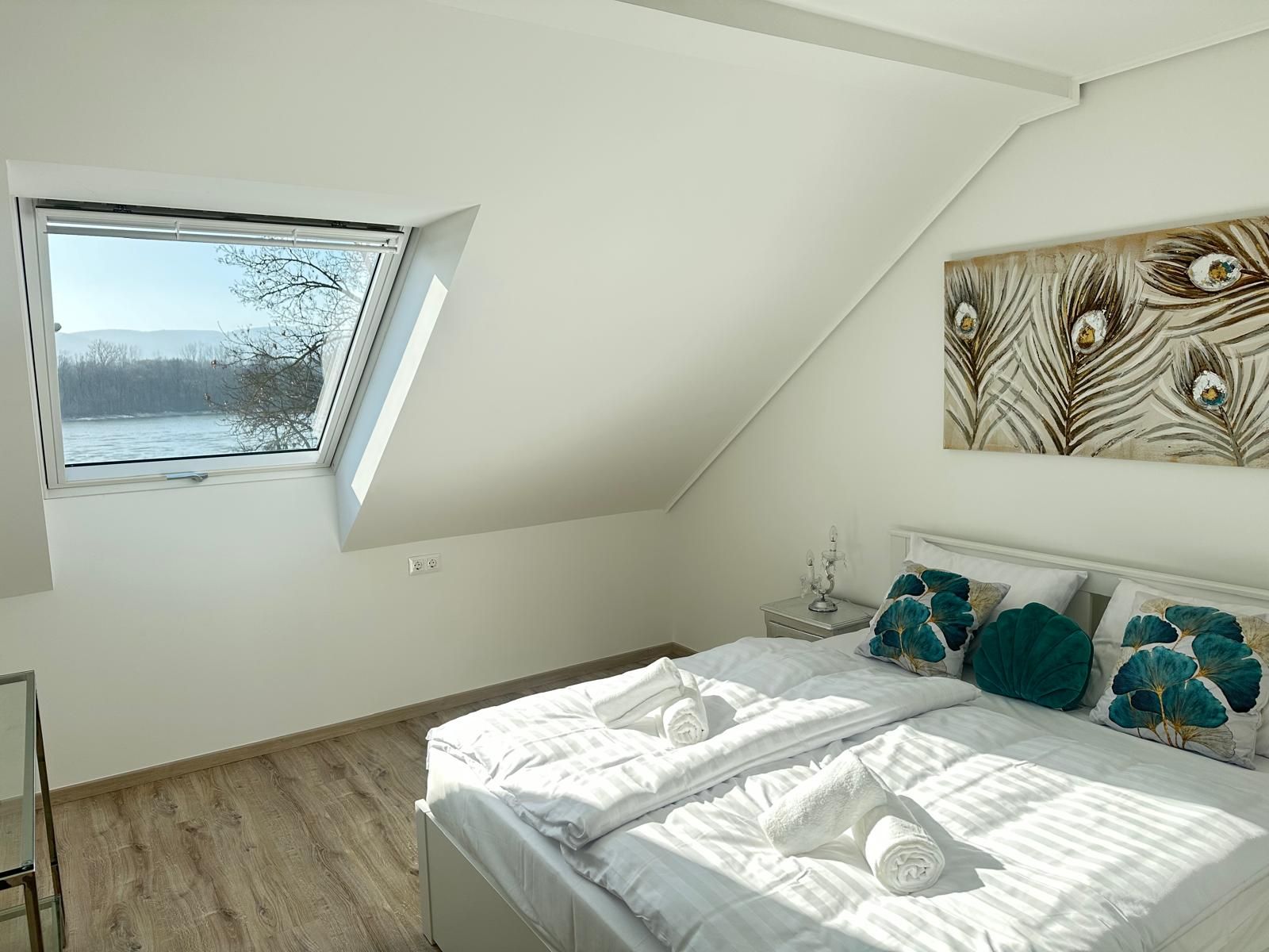 Une chambre dotée d’une lucarne, d’un lit avec du linge de lit blanc, d’oreillers turquoise et d’une œuvre d’art accrochée au mur.
