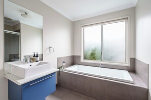 A modern bathroom with a double sink vanity, a bathtub, and a large window with a view of greenery. - Oakdean Coastal Escape | Pet Friendly (Ocean Grove)