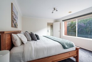 A bedroom with a wooden bed, white bedding, and a view of greenery through a window. - Oakdean Coastal Escape | Pet Friendly (Ocean Grove)
