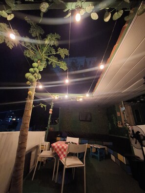 An outdoor dining area with a table and chairs, a hanging plant with green leaves, and string lights.