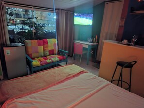 A hotel room with a bed, a colorful sofa, a desk, a chair, a refrigerator, and a TV.
