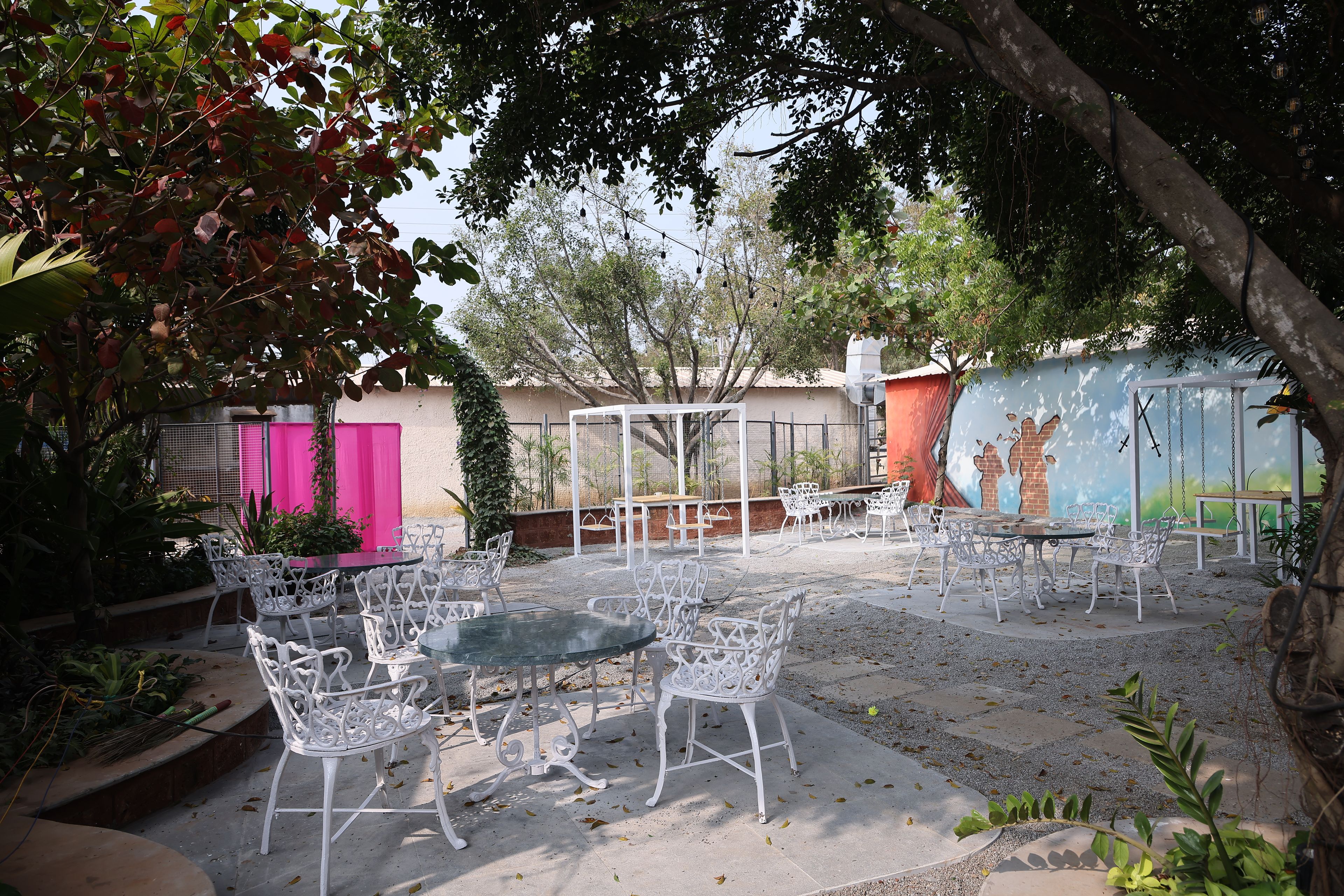 Une terrasse extérieure avec des chaises et des tables en métal blanc, entourée d’arbres et d’un bâtiment orné d’une fresque murale.