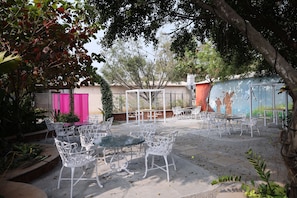 Un patio al aire libre con sillas y mesas de metal blanco, rodeado de árboles y un edificio con un mural.