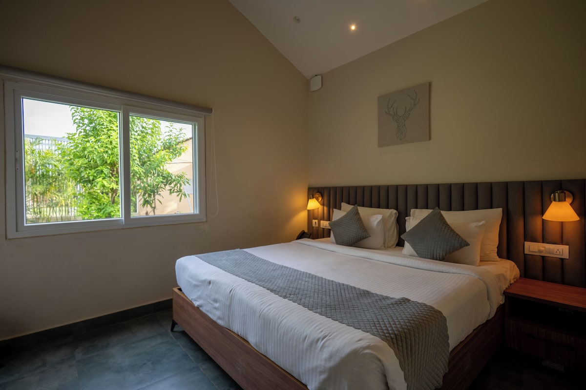 A hotel room with a large bed, two bedside lamps, a window with a view of greenery, and a wall-mounted artwork.