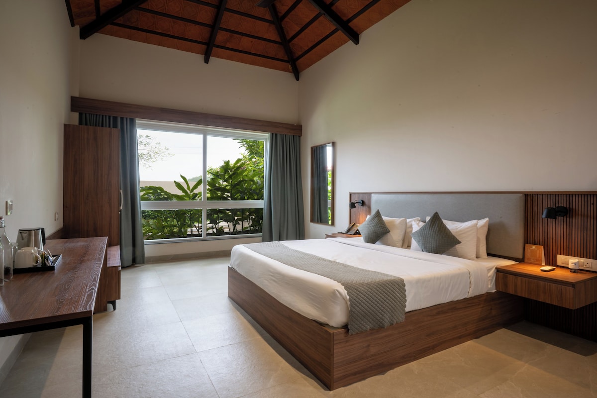 A modern hotel room with a large bed, a wooden desk, a nightstand, and a view of greenery through the window.