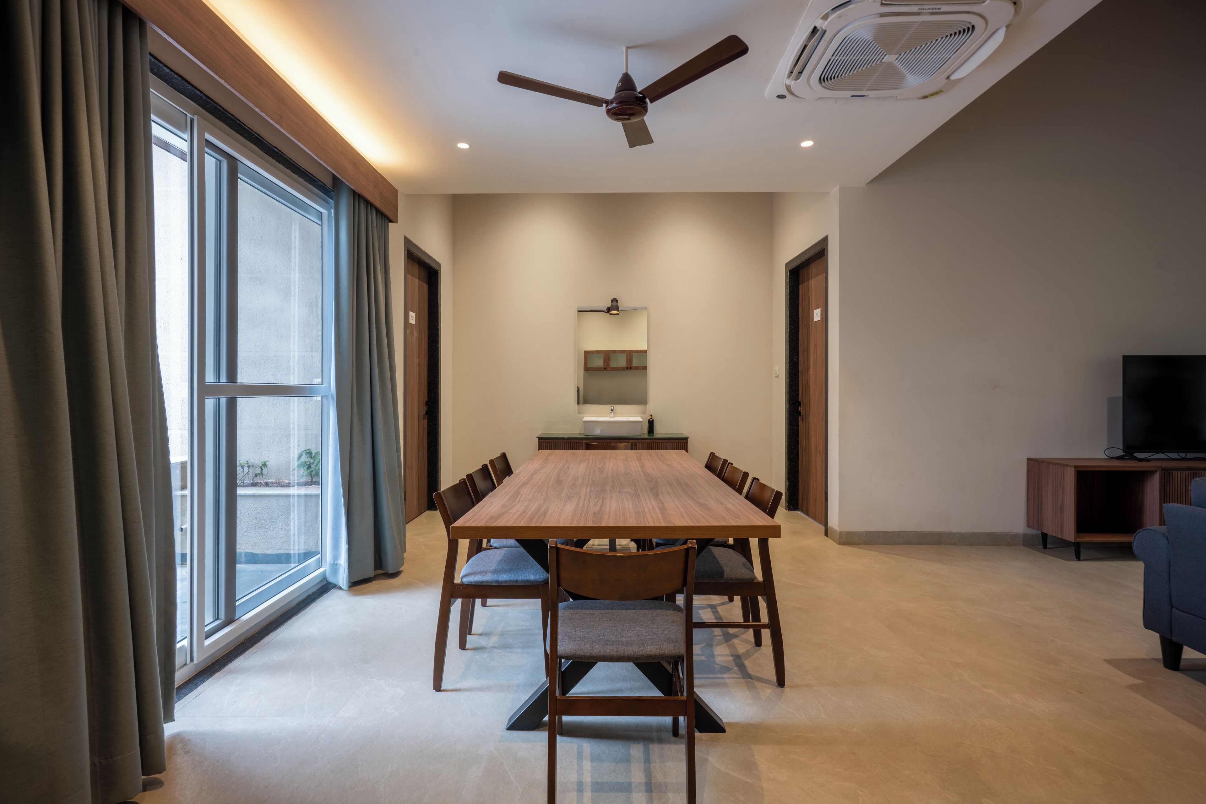 A spacious room with a large dining table, chairs, a ceiling fan, a TV, and a large window with curtains.