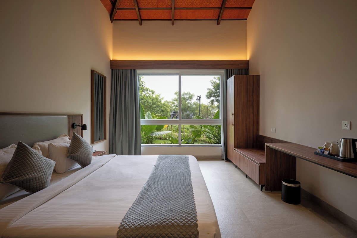 A modern hotel room with a large bed, a desk, and a view of greenery outside.