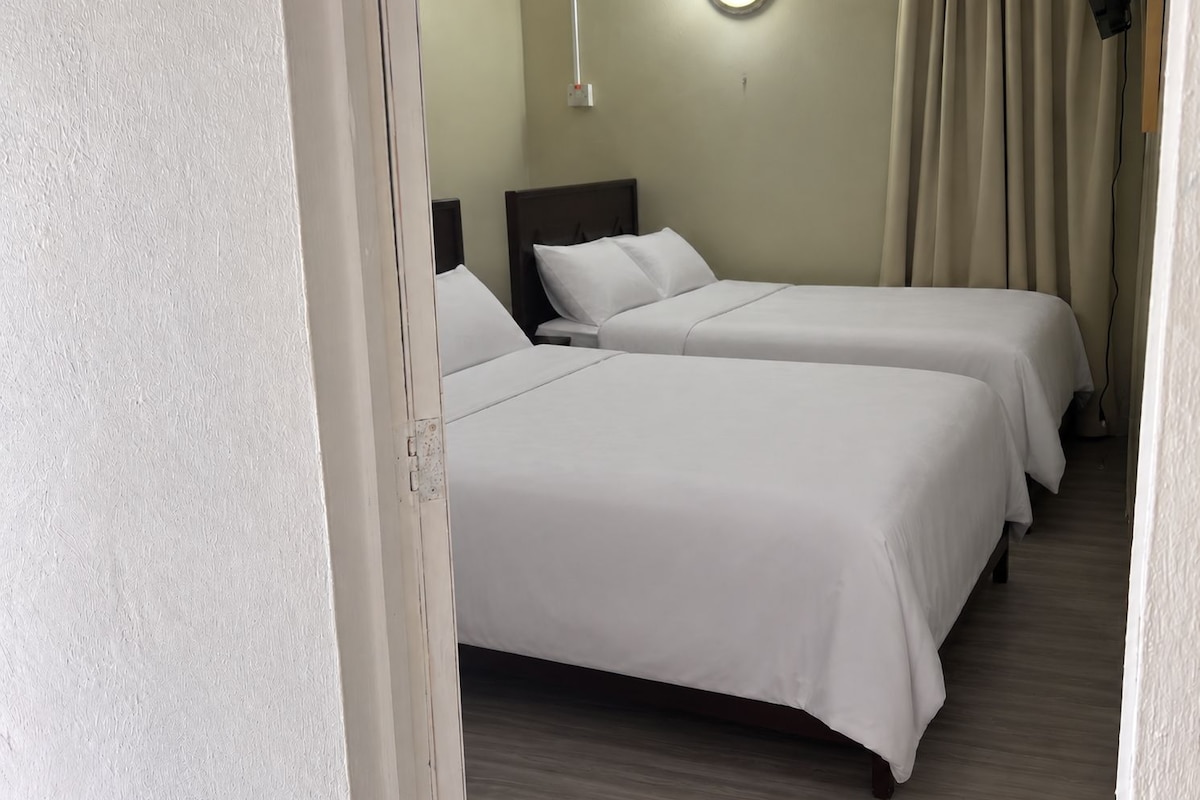 Hotellihuone, jossa on kaksi erillistä sänkyä, yöpöytä, seinään kiinnitetty valaisin ja verho.