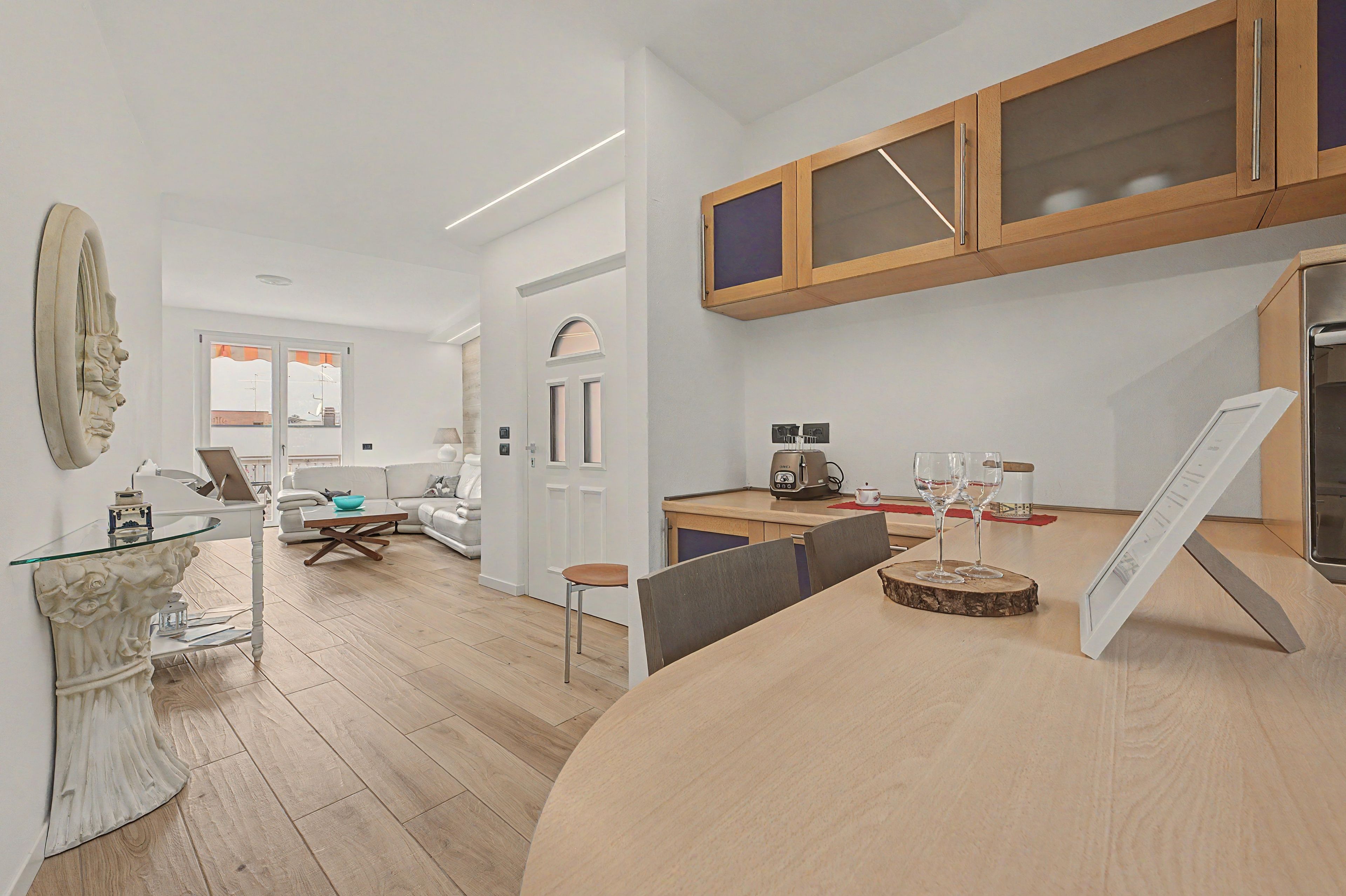 Une cuisine moderne avec un plan de travail en bois, un coin repas doté d’une table en verre et un salon avec un canapé et un élément décoratif mural.