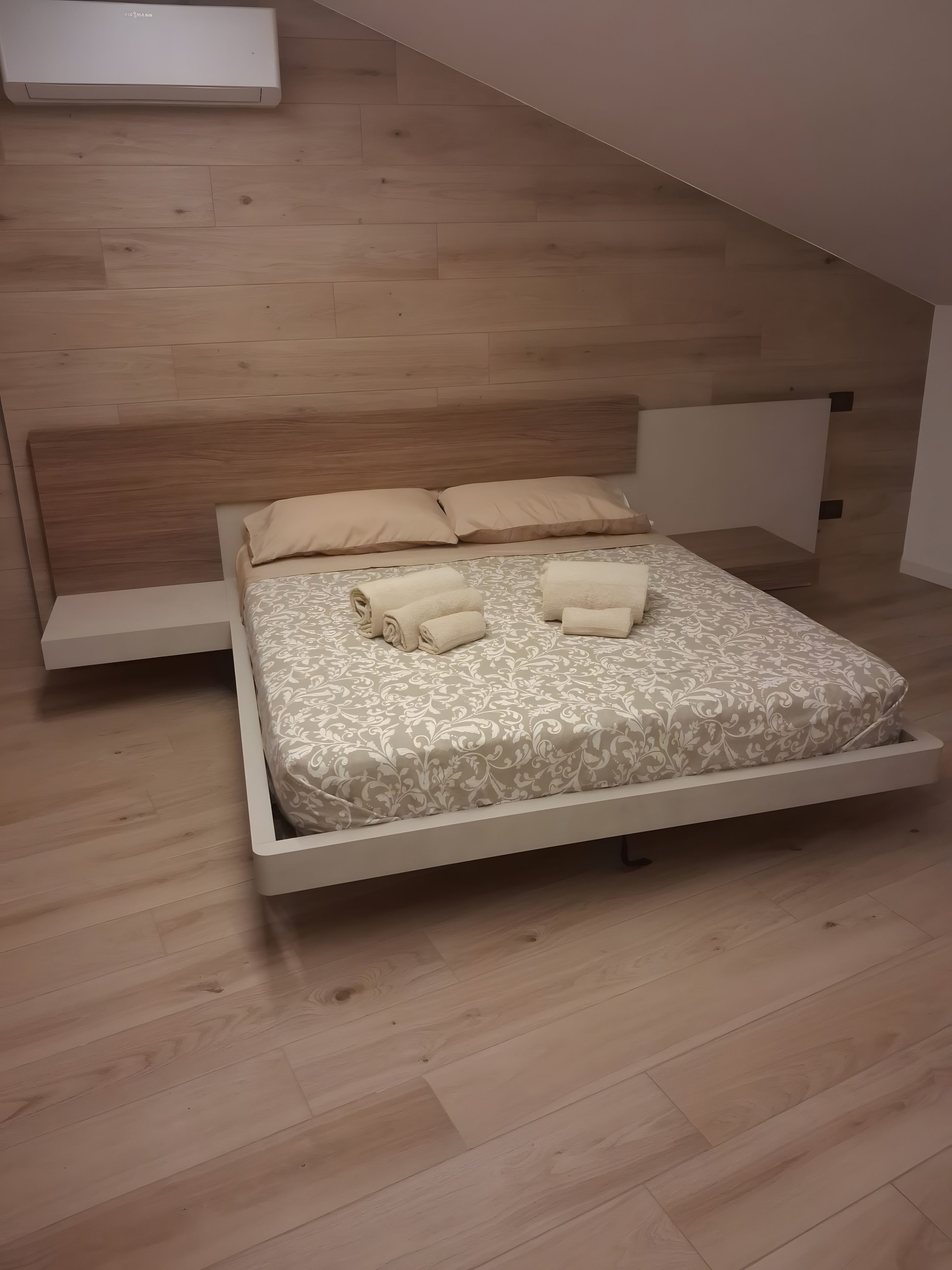 Een moderne slaapkamer met een houten vloer, een bed met een bedovertrek met een patroon, twee kussens en twee opgerolde handdoeken.
