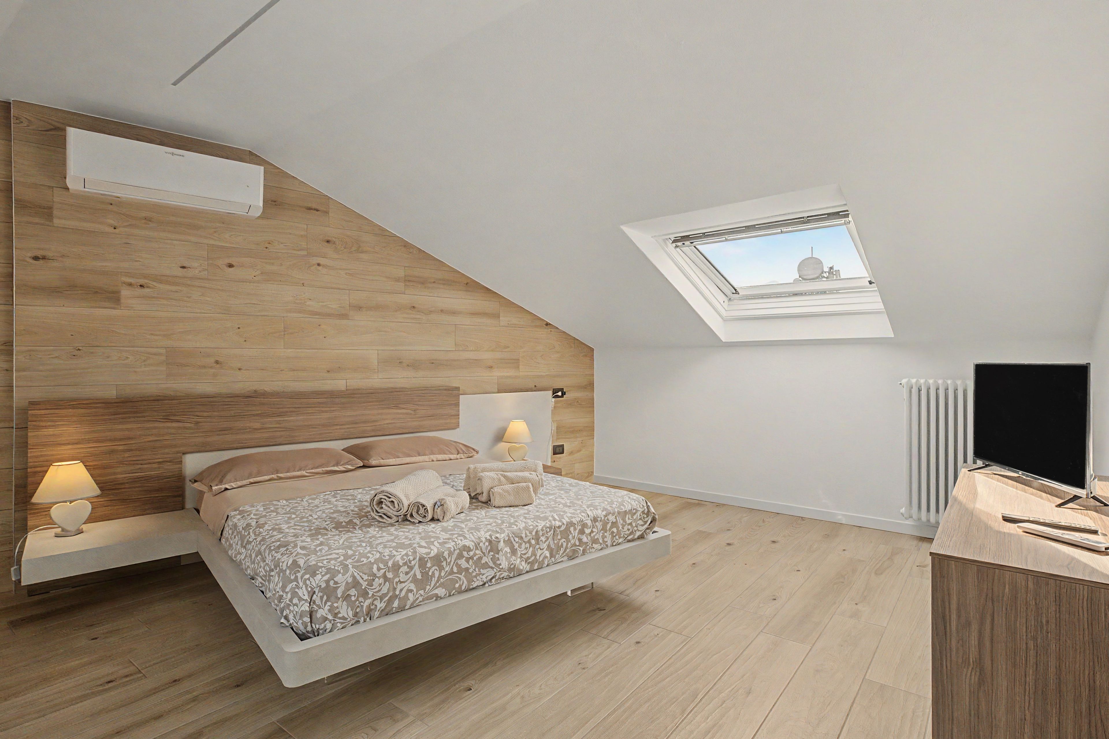Ein Schlafzimmer mit schrägverdeckter Decke, einem Bett mit Kopfteil, einem Nachttisch mit Lampe, einem Heizkörper und einem Fernseher.