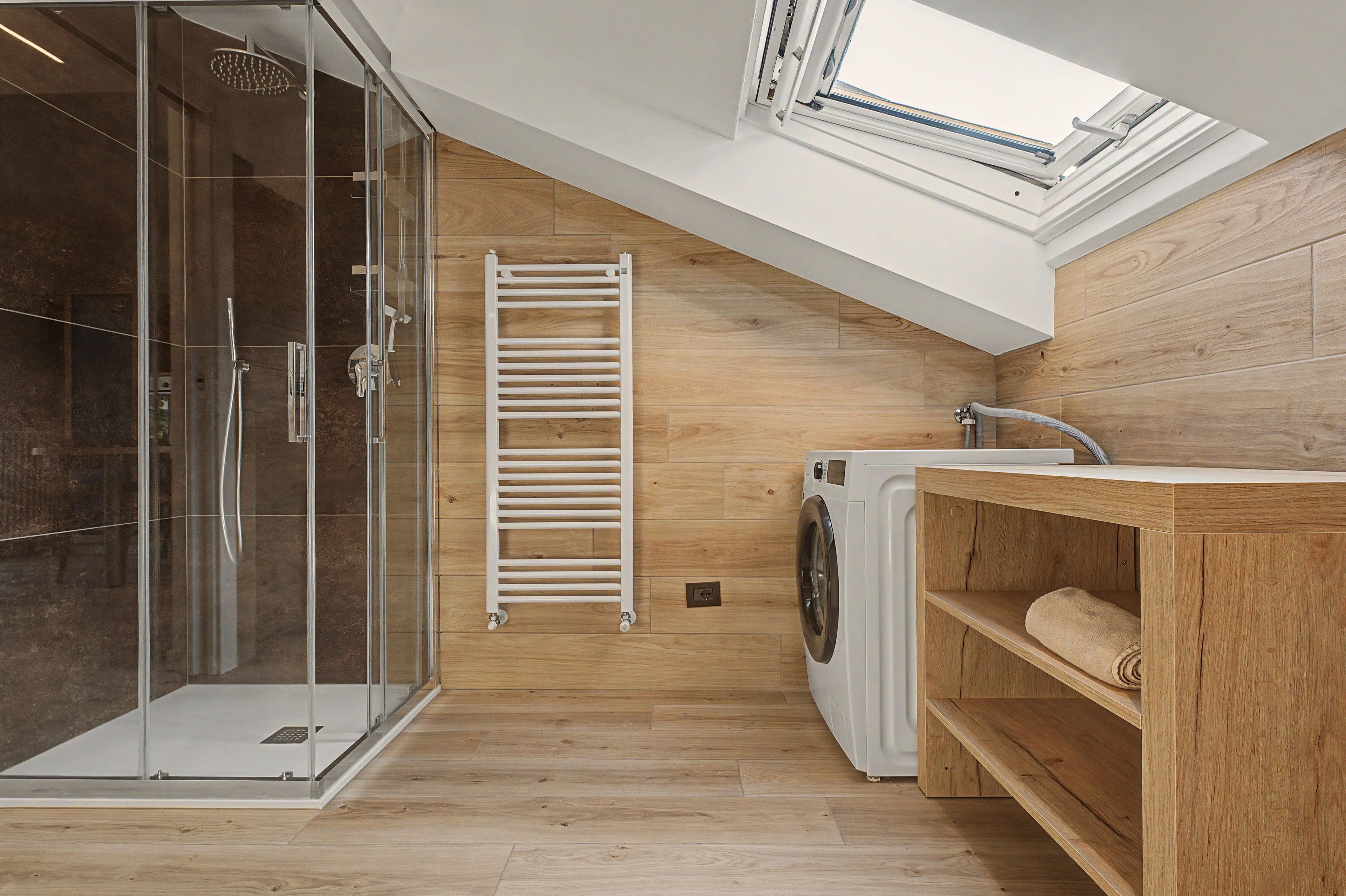 Une salle de bain moderne avec une douche à parois de verre, un plancher en bois, un lave-linge encastré et une lucarne.