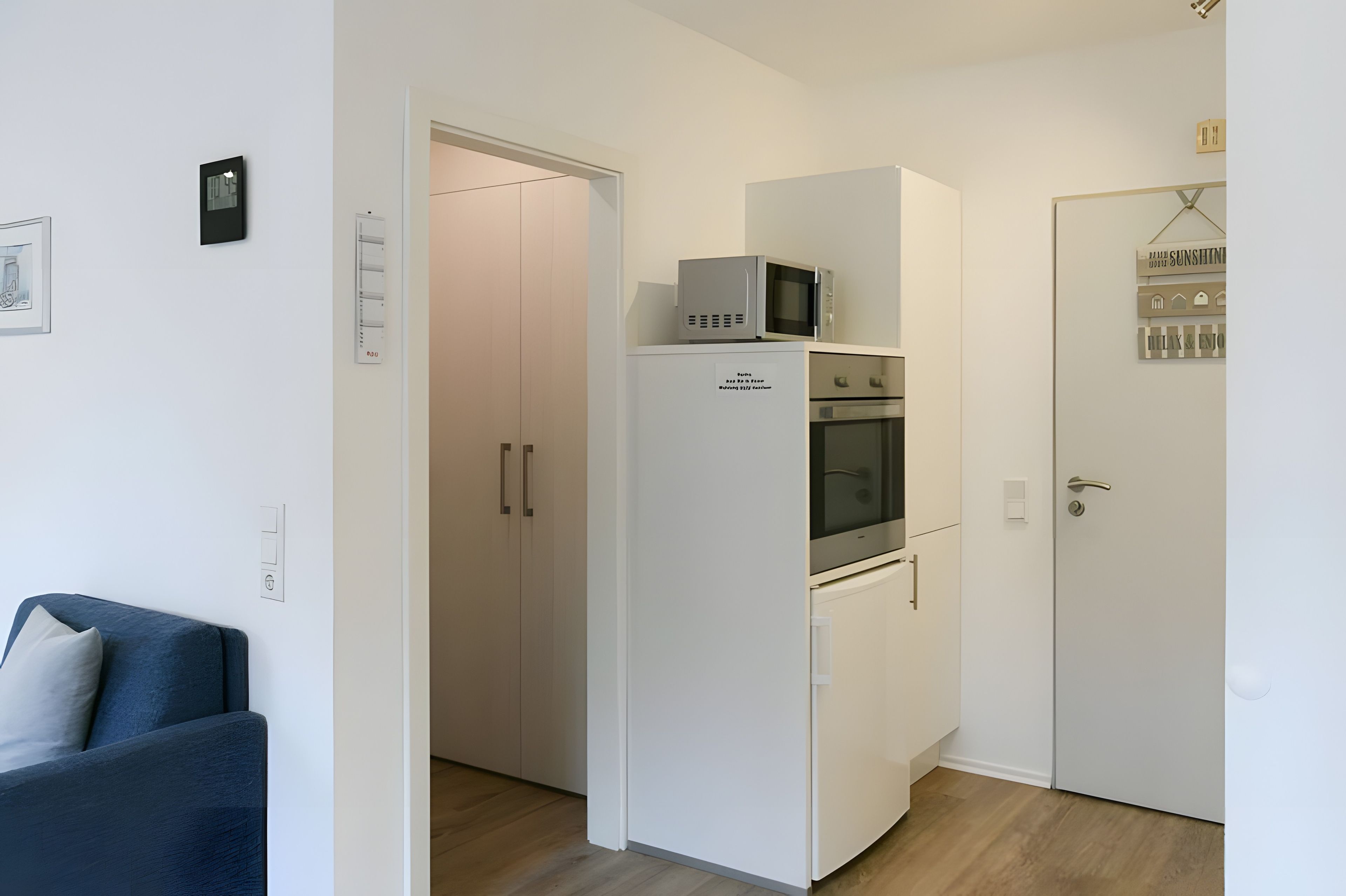Eine kompakte Küche mit Mikrowelle, Backofen und kleinem Kühlschrank. Eine Tür mit einem Schild darüber und ein Lichtschalter an der Wand sind vorhanden.