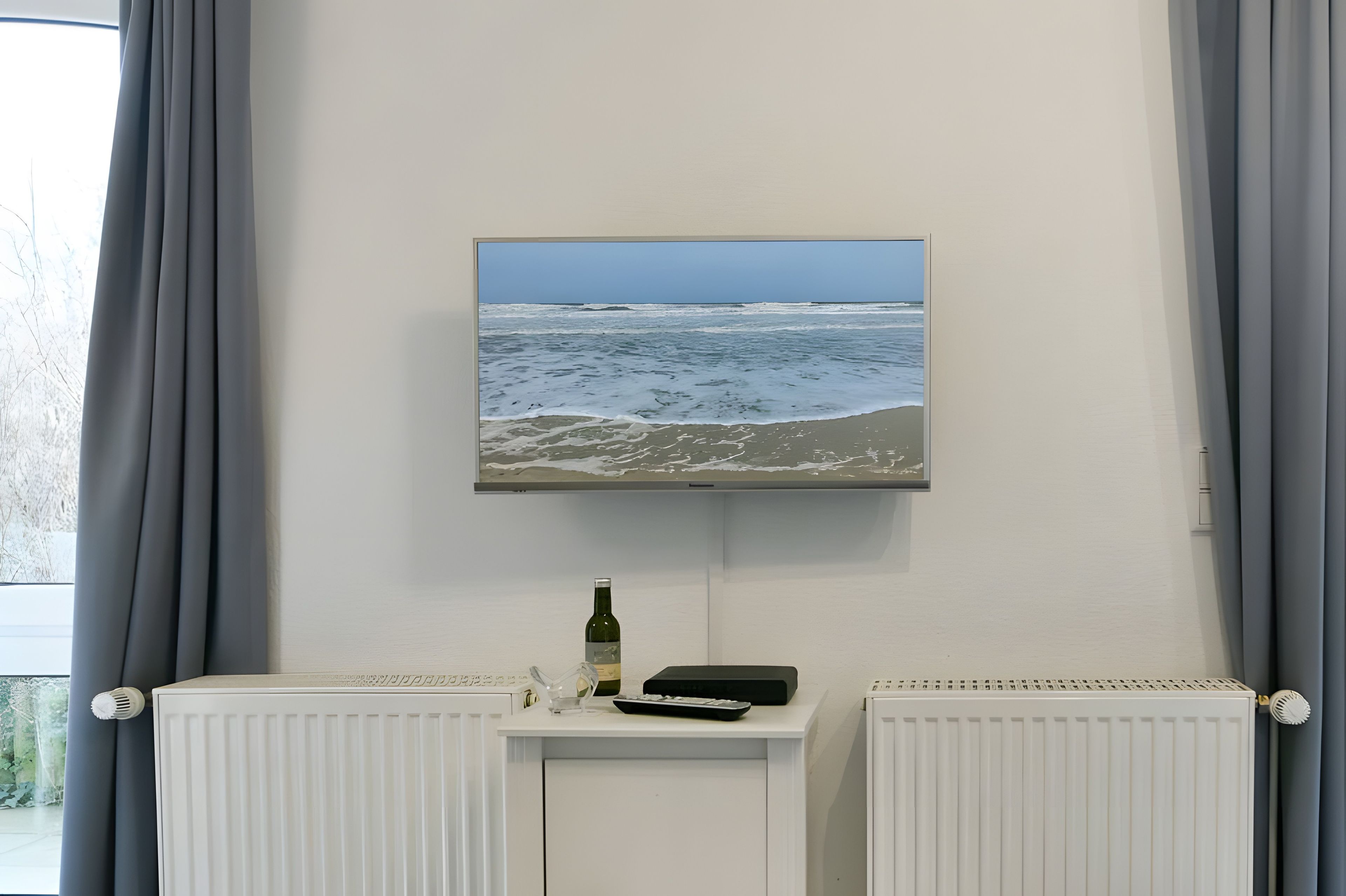 Un téléviseur fixé au mur affiche une scène de plage. Une bouteille et une télécommande sont posées sur une petite table en dessous.