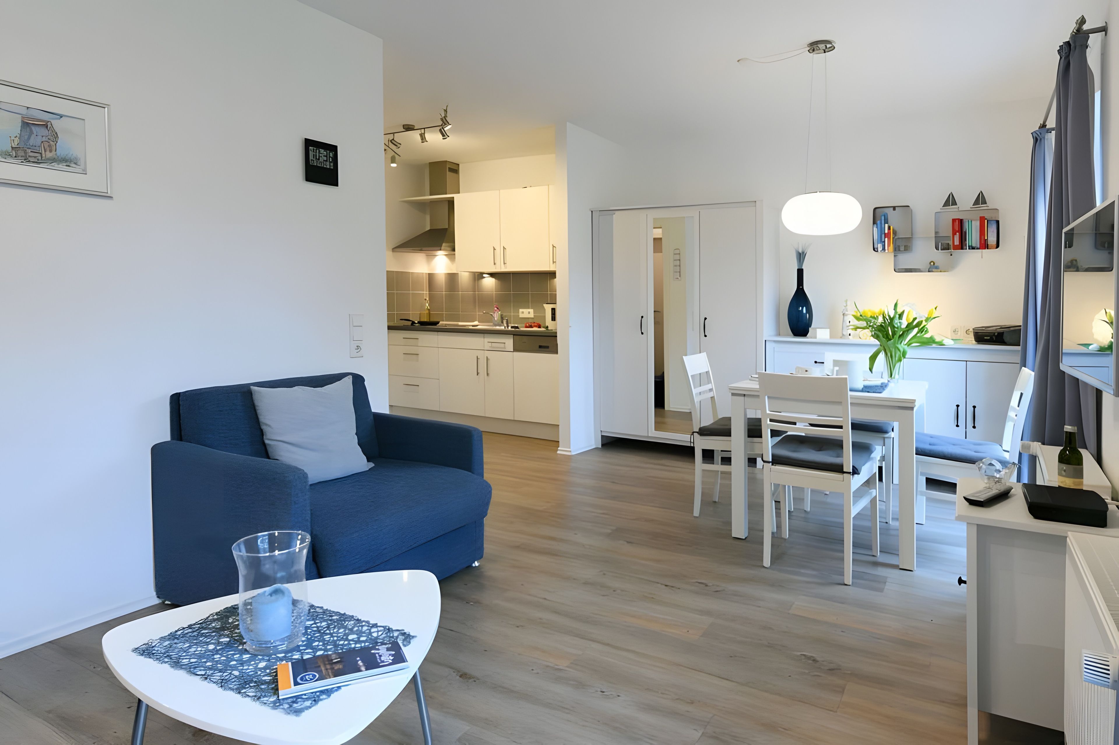 Ein modernes Wohnzimmer mit einem blauen Sofa, einem Esstisch mit Stühlen und einer Küchenzeile mit weißen Schränken und einer Pendelleuchte.