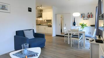 Ein modernes Wohnzimmer mit einem blauen Sofa, einem Esstisch mit Stühlen und einer Küchenzeile mit weißen Schränken und einer Pendelleuchte.