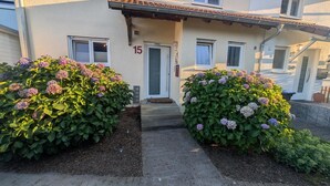 Um edifício com entrada numerada 15, varanda coberta e dois grandes arbustos com flores de hortênsia.