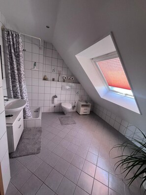 Ein modernes Badezimmer mit Oberlicht, weißen Fliesen, einer Dusche, einer Toilette und einem Waschbecken.
