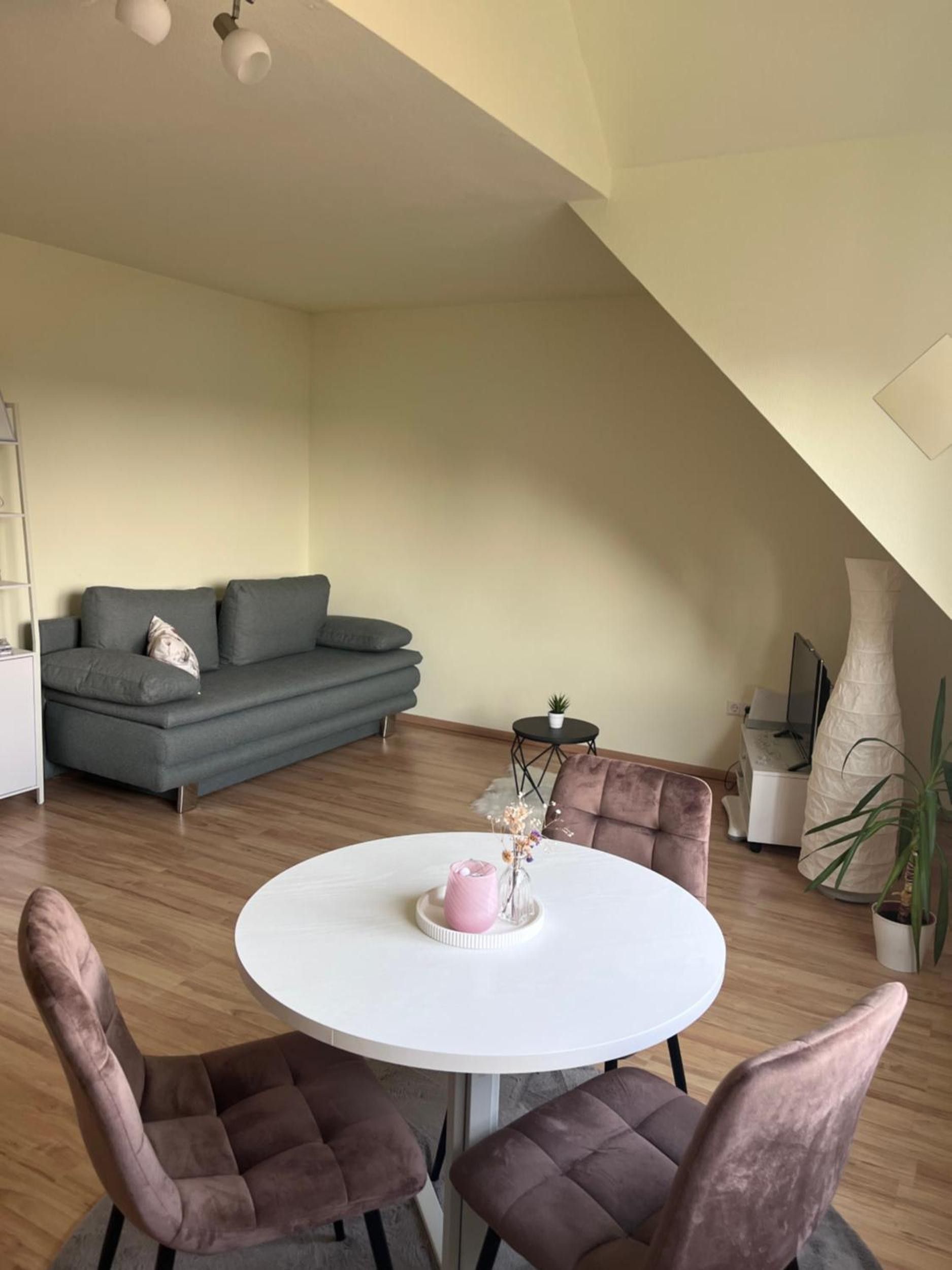 Ein modernes Wohnzimmer mit einem grauen Sofa, einem runden Esstisch mit Stühlen und einem kleinen Beistelltisch mit einer Pflanze.